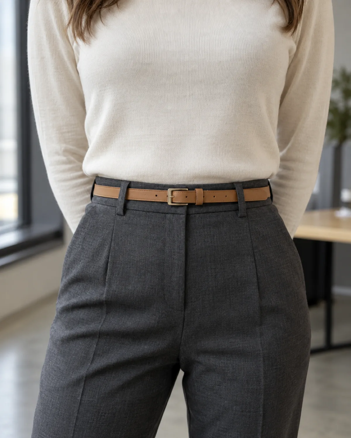 A close crop on the waist showing a cream fine-gauge knit tucked into dark grey high-waisted trousers with a slim camel leather belt threaded through the belt loops, plain metal buckle visible