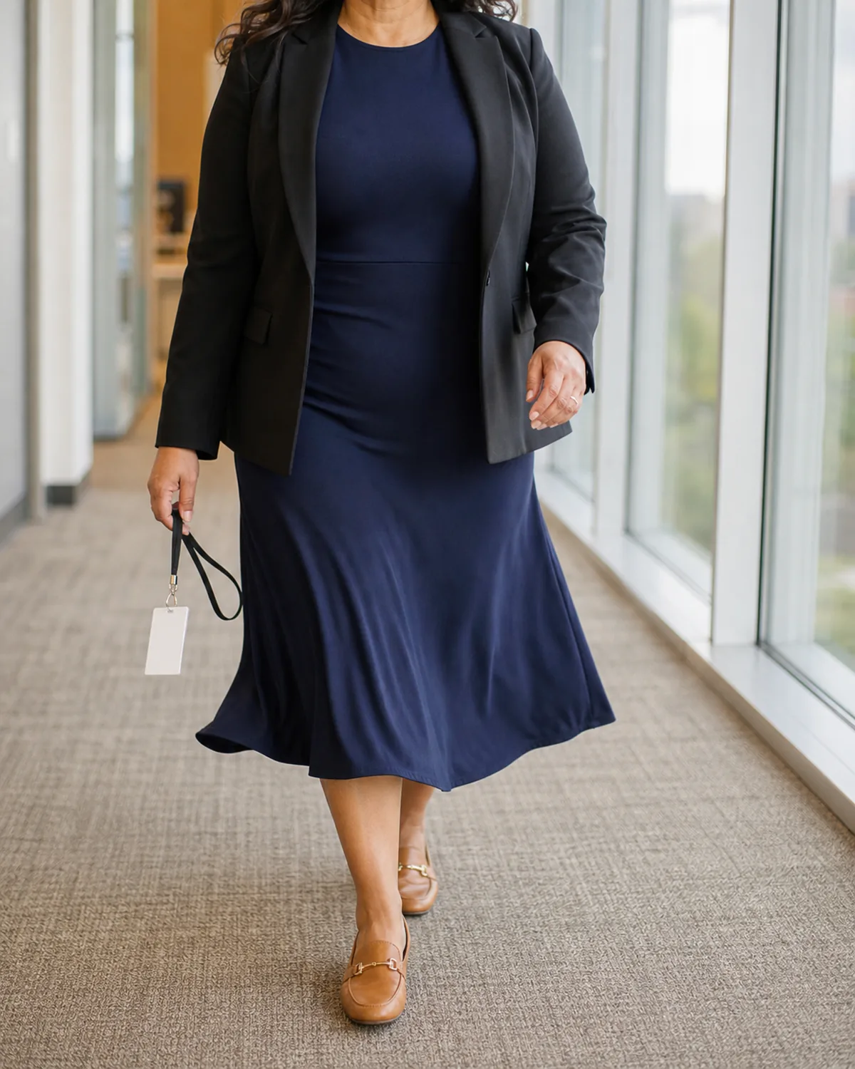 A curvy woman walking mid-stride in a black blazer over a navy A-line midi dress with camel loafers, holding a lanyard badge, in a bright office corridor