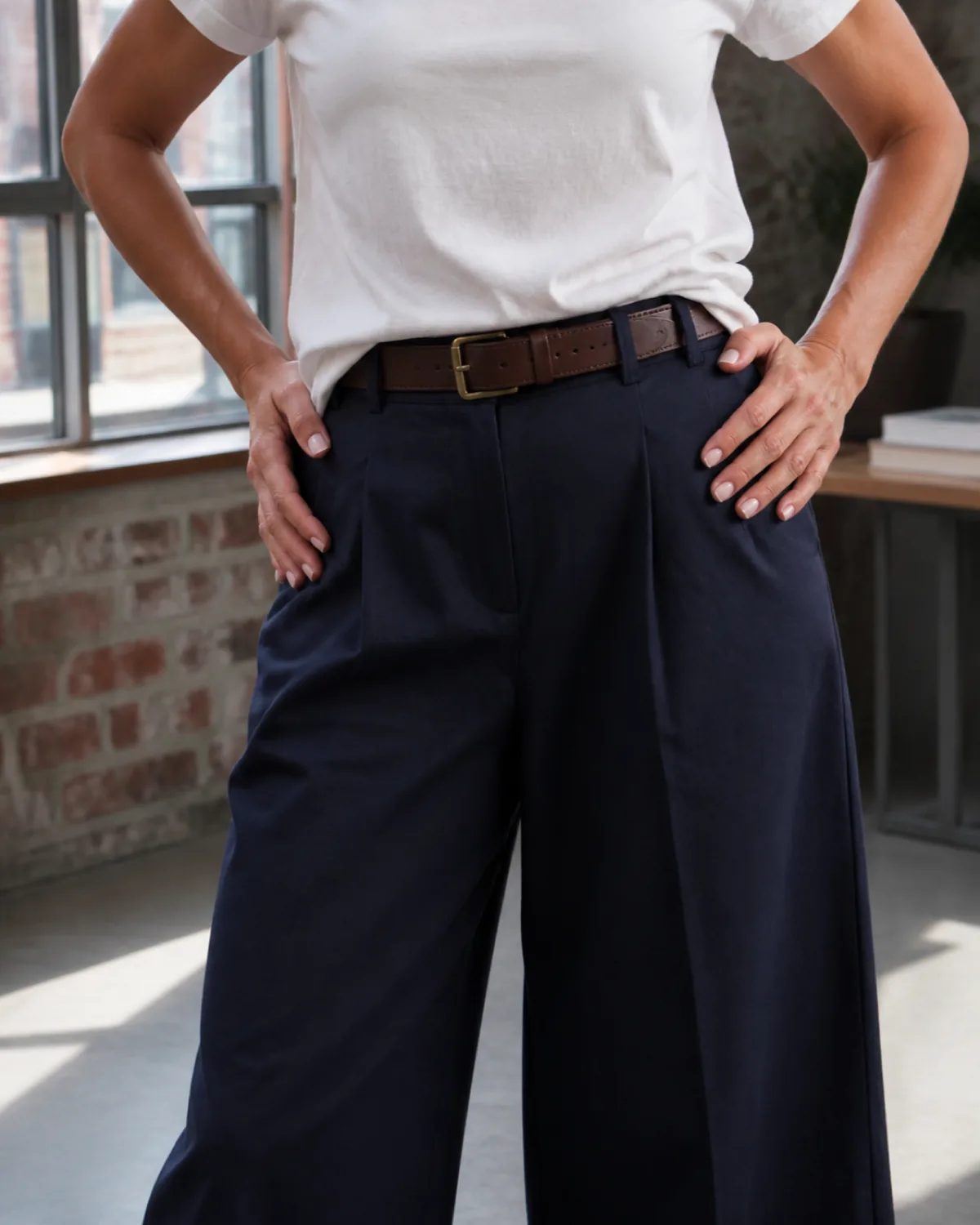 A close crop of navy pressed culottes with a tucked white crew-neck tee and brown leather belt, hands in the pockets, worn with tan wide-band leather slides in a brick-walled studio office