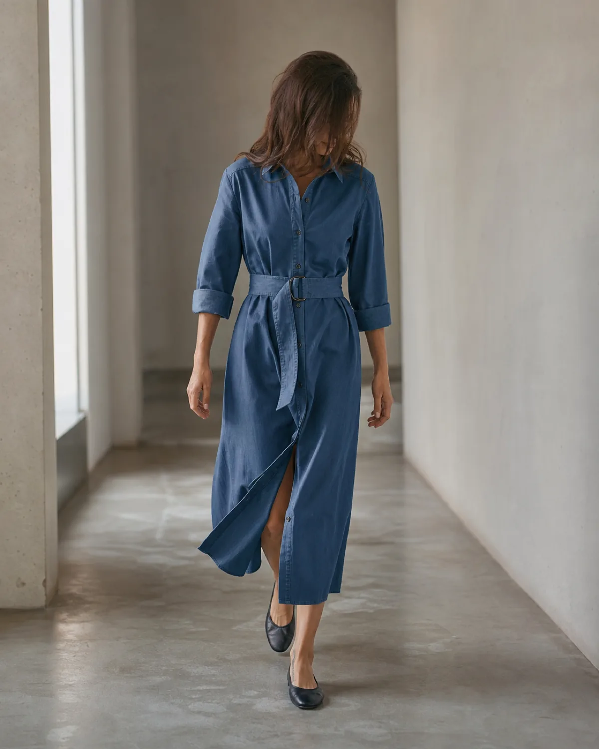 A woman walking along a plain corridor in a mid-blue belted cotton shirtdress at midi length with black ballet flats