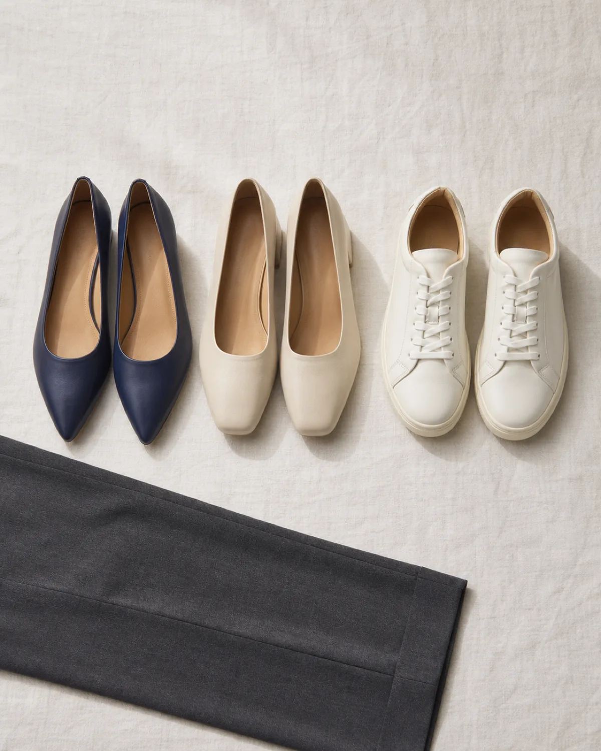Overhead flat lay of three pairs of unbranded shoes on pale linen: navy pointed flats, cream low block-heel court shoes and white leather sneakers, with a folded charcoal trouser leg entering the frame