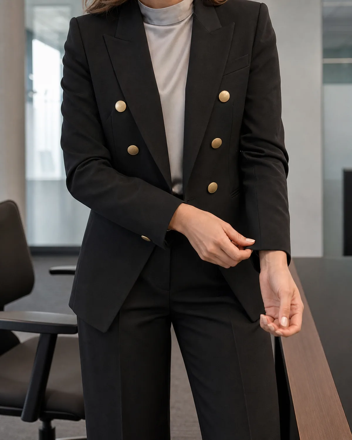 A mid-torso crop of a woman adjusting her cuff on a black double-breasted blazer with gold-tone buttons, wearing a pale grey silk mock-neck shell and high-waisted black trousers