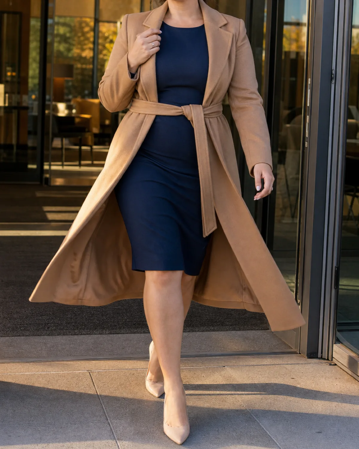 A midsize woman walking in a camel wrap coat belted at the waist over a navy sheath dress with nude pumps