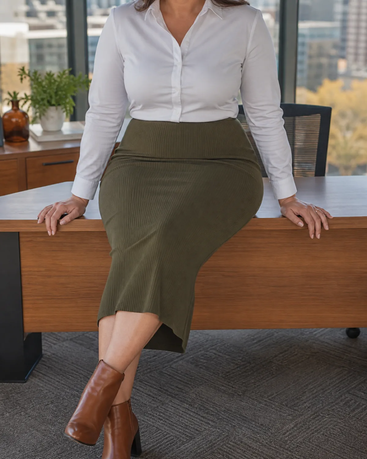 A midsize woman seated at a desk edge in an olive ribbed knit midi skirt with a white button-down and cognac ankle boots