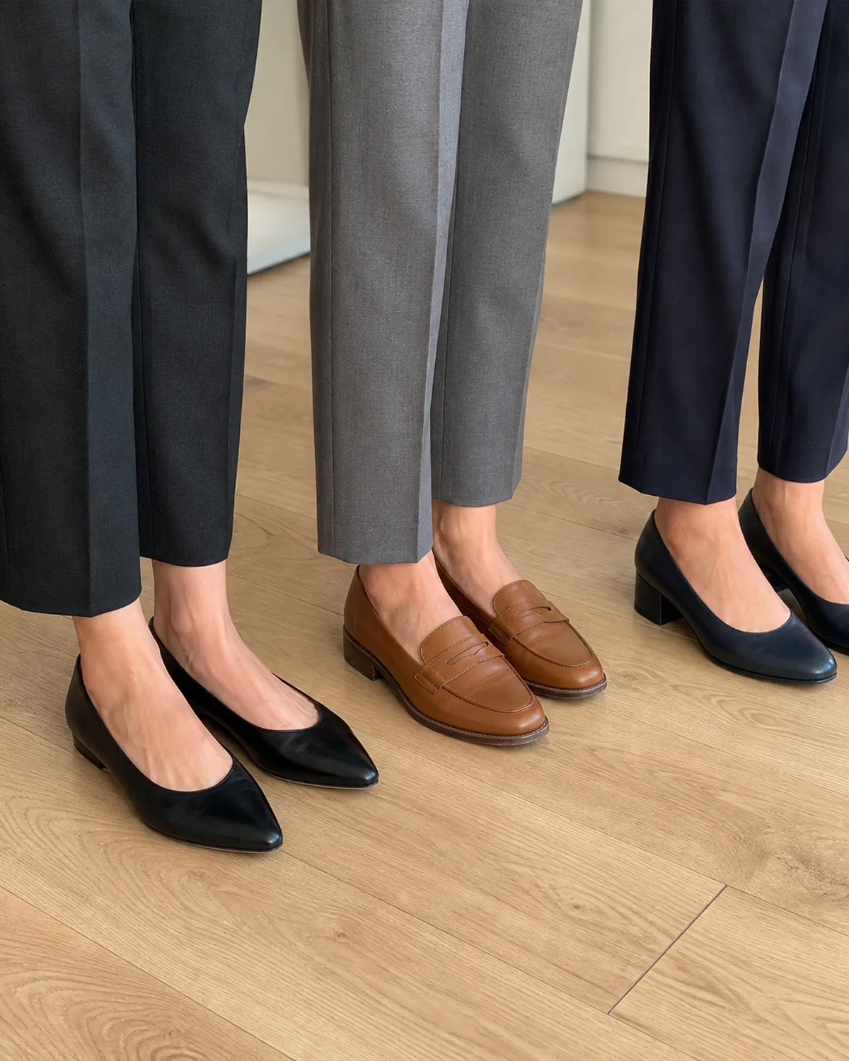 Three pairs of professional shoes on a light oak floor: black pointed-toe flats with dark trousers, tan penny loafers with grey trousers, and navy block-heel pumps with navy trousers