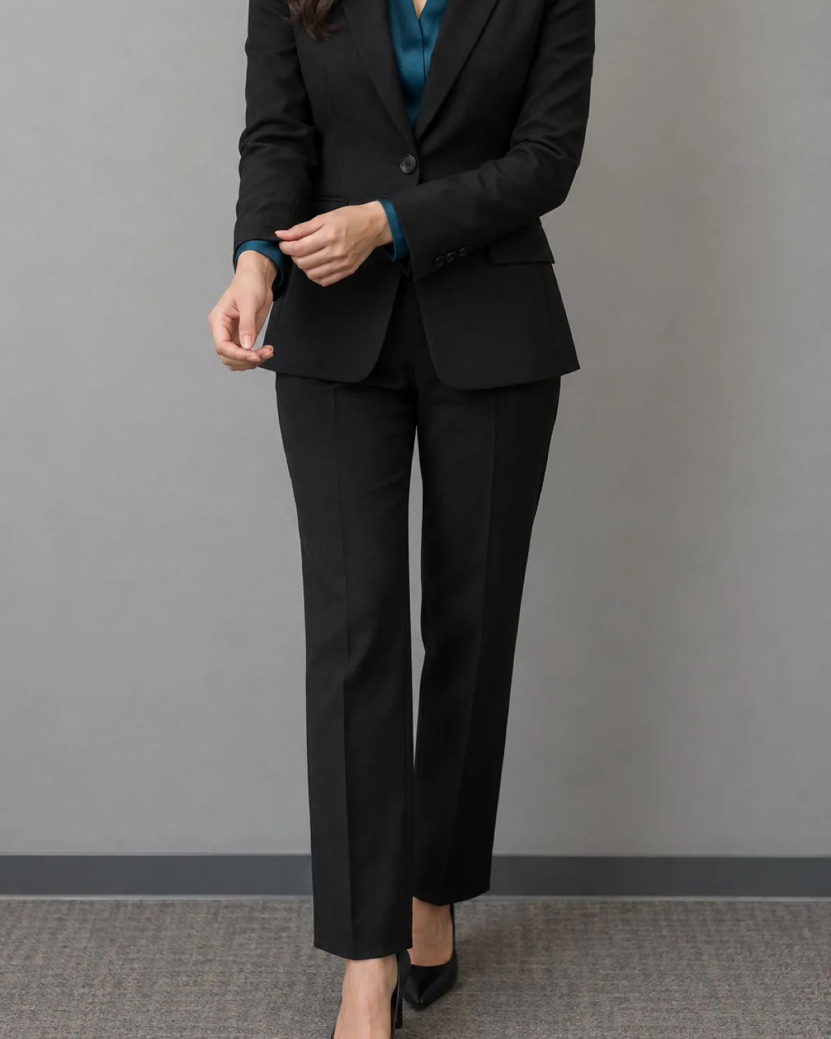 A woman in a mid-torso crop wearing a black single-breasted wool trouser suit with a deep teal silk blouse visible at the blazer's V, adjusting one blazer cuff with the other hand