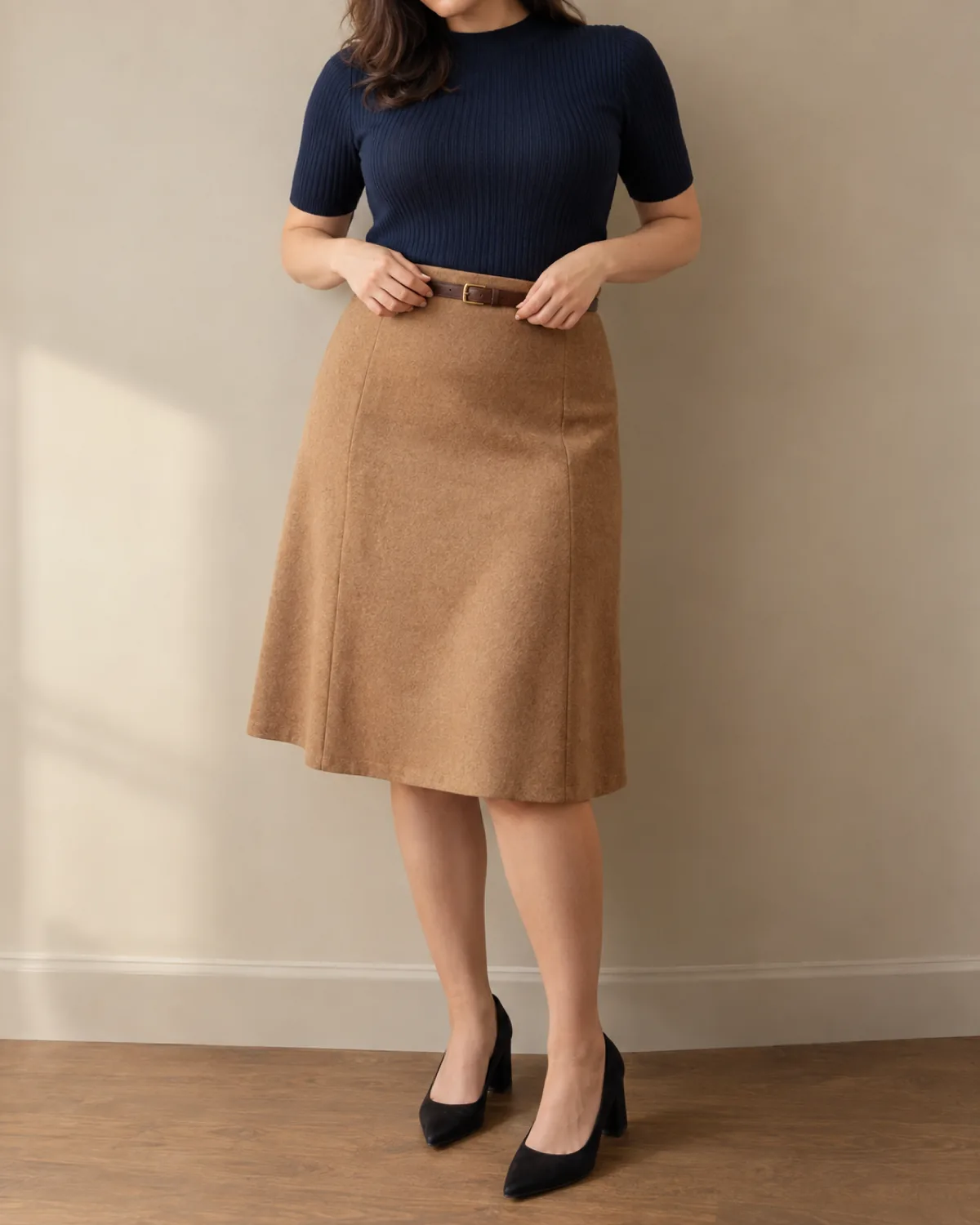 A midsize woman adjusting a thin leather belt at her waist, wearing a navy ribbed knit tucked into a camel A-line skirt