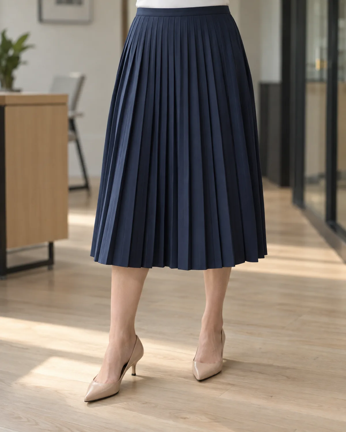 Lower half of a midsize woman wearing a navy pleated midi skirt with nude kitten heels, the edge of a white tee visible at the waist