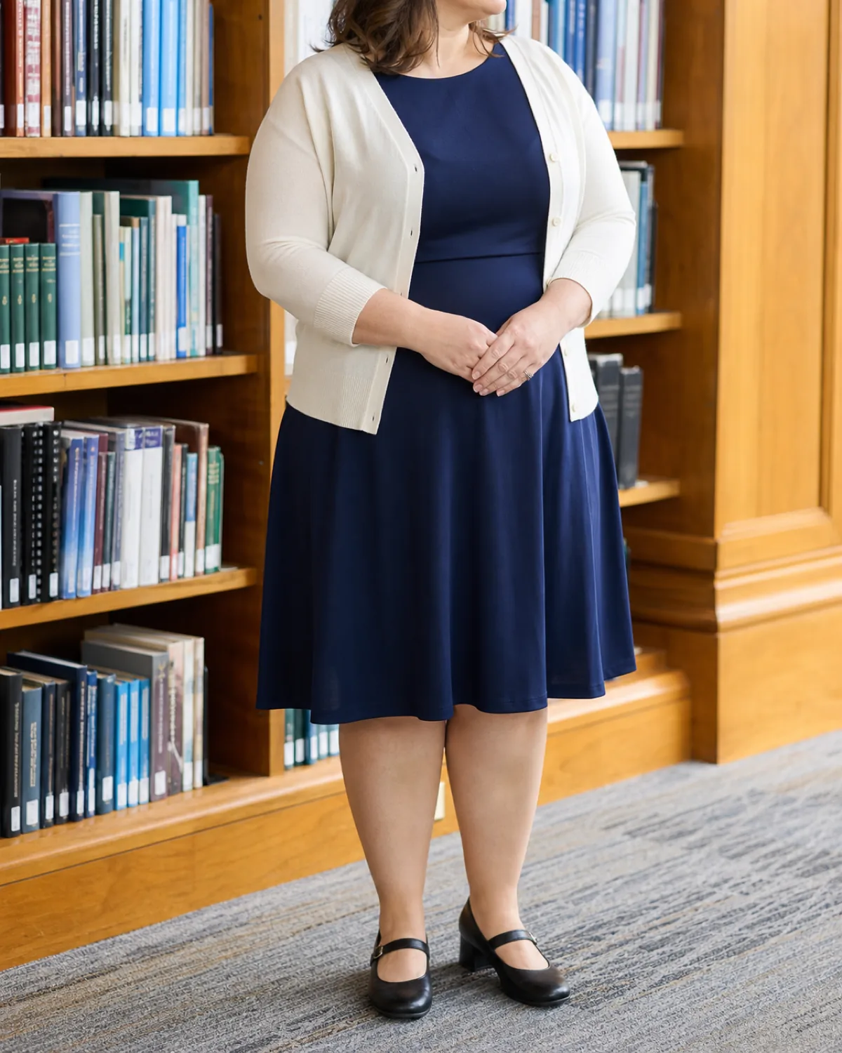 A plus-size woman standing in front of library bookshelves in a navy fit-and-flare dress with a knee-length hem and a cream fine-knit cardigan, hands clasped at waist, wearing black Mary Jane shoes