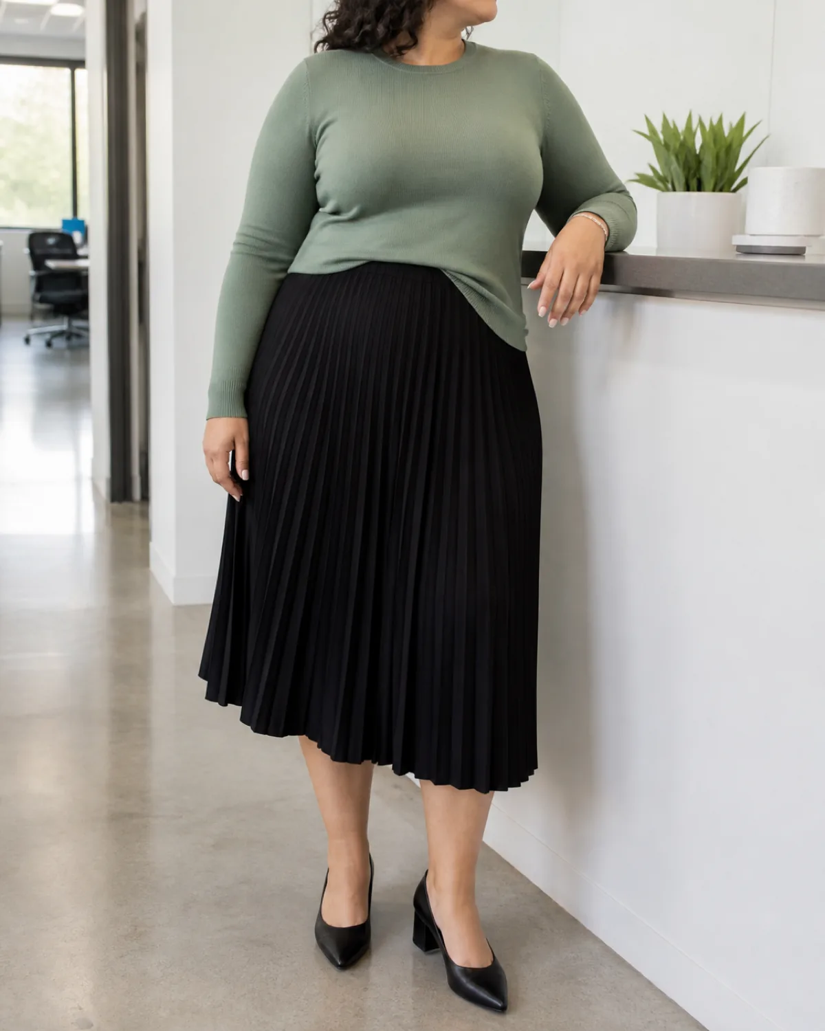 A plus-size woman leaning slightly against a reception counter in a retail office, wearing a sage green fine-gauge knit top and a black pleated midi skirt with black block heels