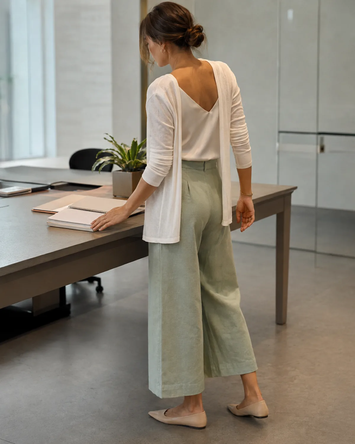 A woman seen from behind at a desk, wearing sage wide-leg cropped trousers, an ivory camisole, and a cream cardigan