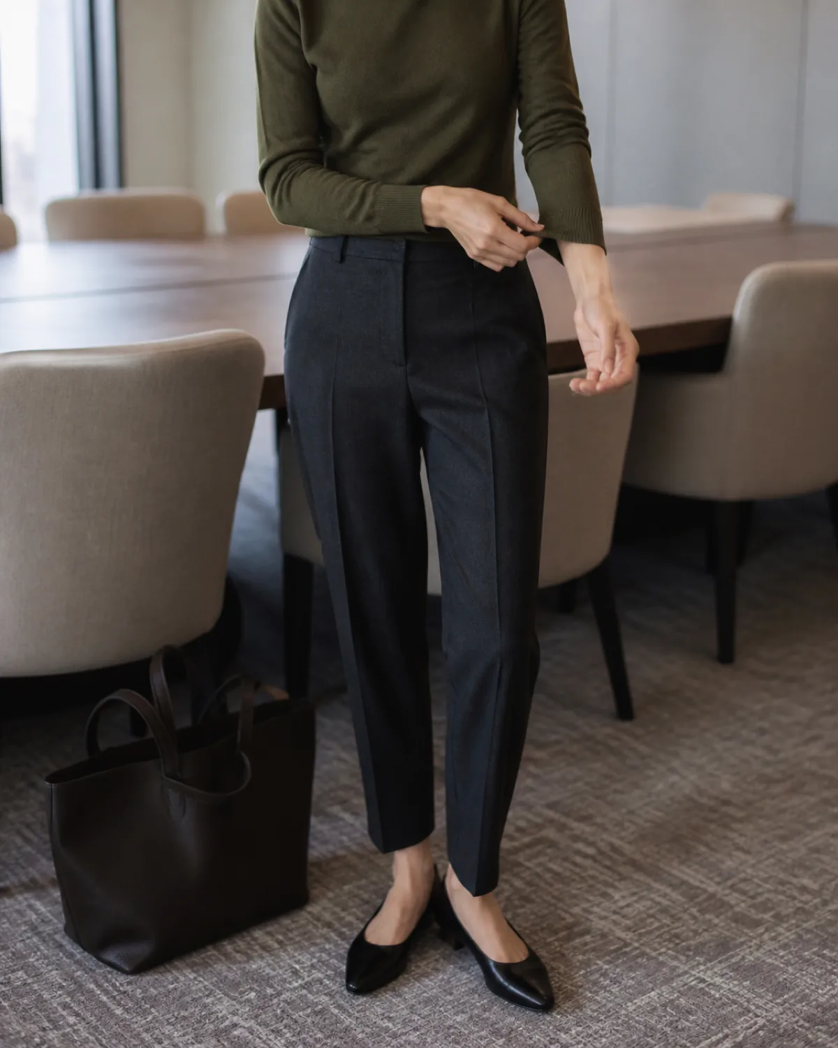 A woman in an olive fine-gauge crewneck sweater and dark charcoal pressed trousers adjusting her cuff in a conference room with upholstered chairs and a dark leather tote on the floor