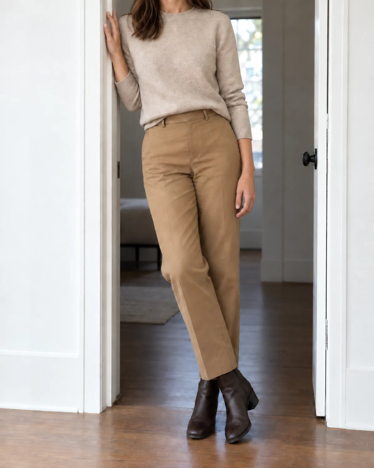 A woman standing in a white doorway leaning one shoulder against the frame, wearing an oatmeal crewneck sweater half-tucked into camel straight-leg trousers and dark brown leather ankle boots with a low block heel