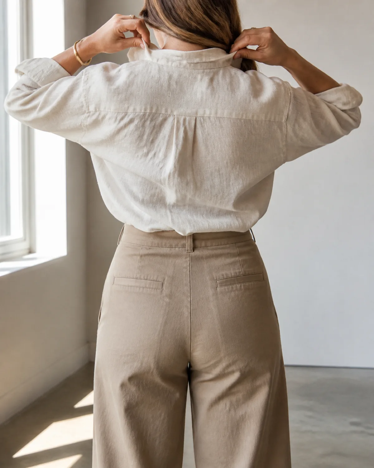 A woman from behind adjusting her collar, wearing a cream linen shirt tucked into tan cotton twill cropped wide-leg trousers ending above the ankle, in a bright studio space