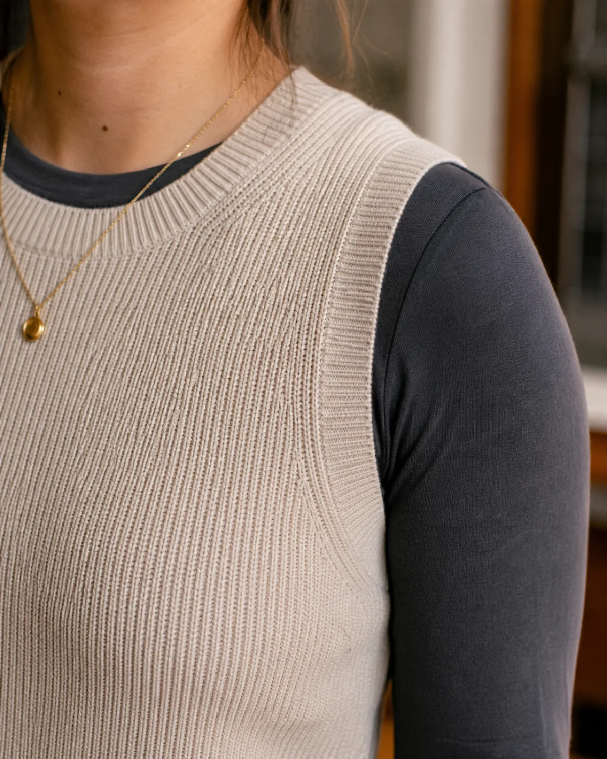 Close-up of an oatmeal ribbed knit vest layered over a charcoal long-sleeve crewneck tee with a fine gold pendant necklace visible, cropped at the shoulder and upper chest