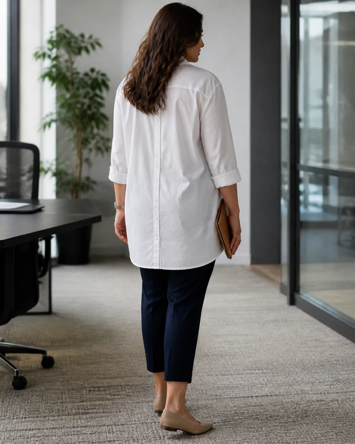 A woman from behind in a white oversized cotton poplin shirt worn as a tunic to mid-thigh with rolled cuffs, navy slim ankle trousers, and tan pointed-toe mules, holding a cognac leather portfolio in one hand in an office