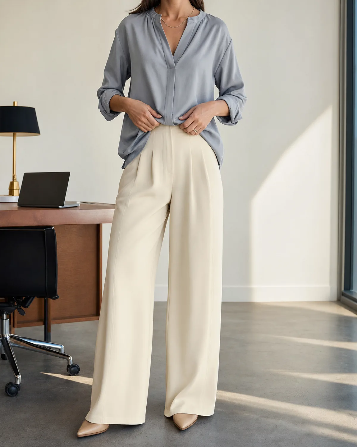 A woman in a mid-torso crop view wearing a grey-blue relaxed blouse tucked loosely at the front into warm ivory wide-leg crepe trousers with a thin chain necklace