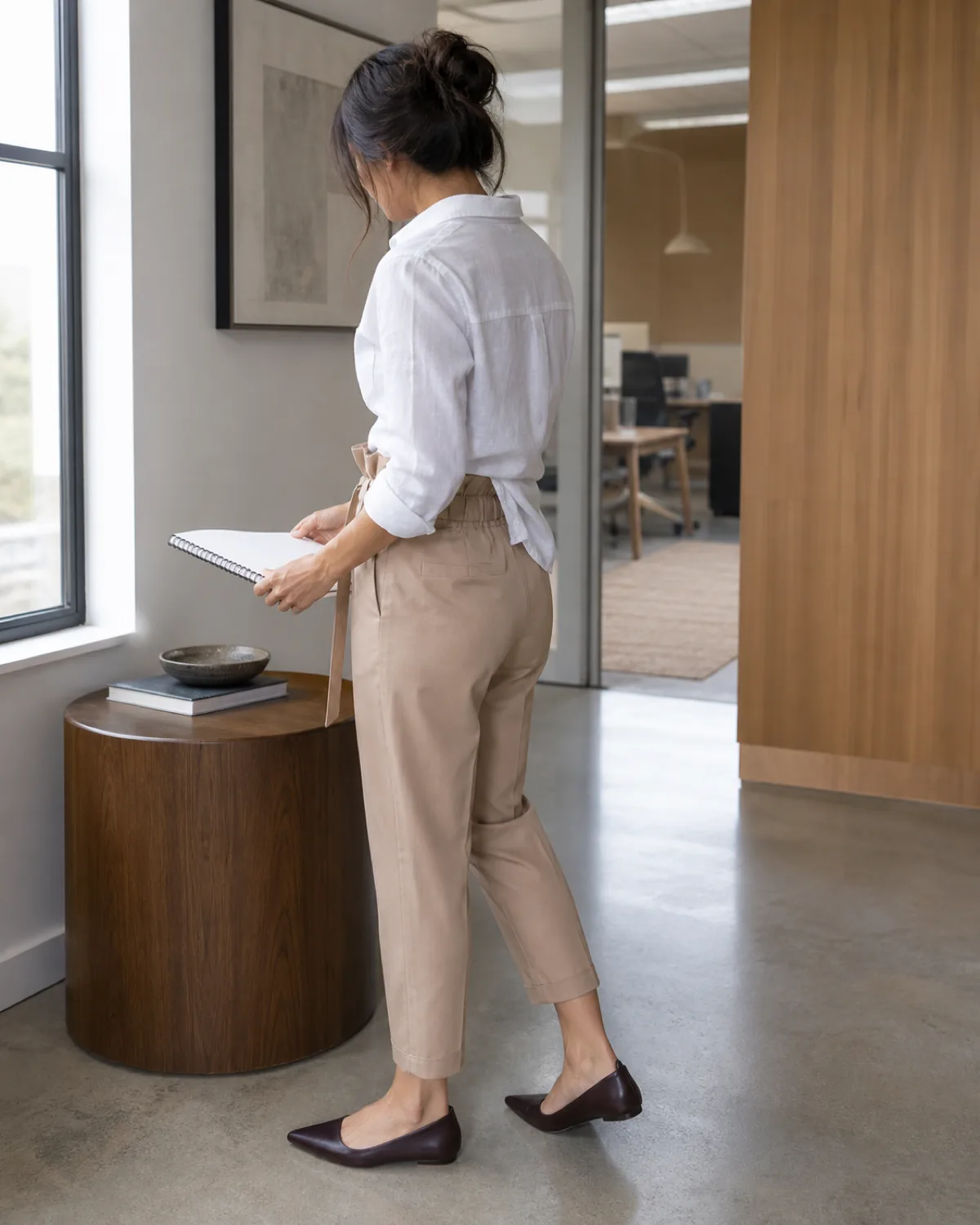 A petite woman in back-three-quarter view looking down at a notebook, wearing tan paper-bag-waist trousers with a self-tie belt and a white linen shirt French-tucked at the front with dark brown pointed-toe flats