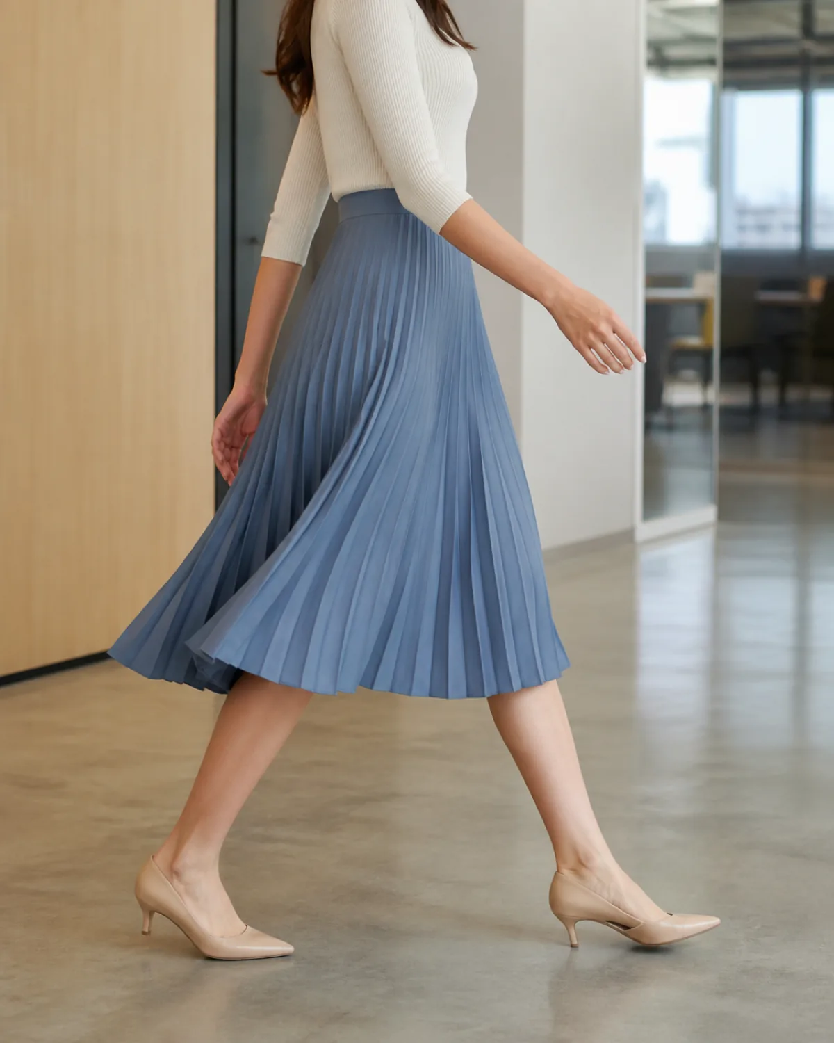 A petite woman walking mid-stride from the side in an office corridor wearing a dusty-blue pleated midi skirt with pleats fanning open, a fitted cream knit top tucked in, and nude kitten heels