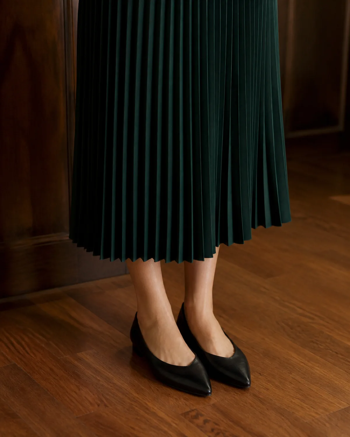 Close crop on a forest-green pleated midi skirt hem falling to mid-calf with black pointed-toe flats on a warm wood floor