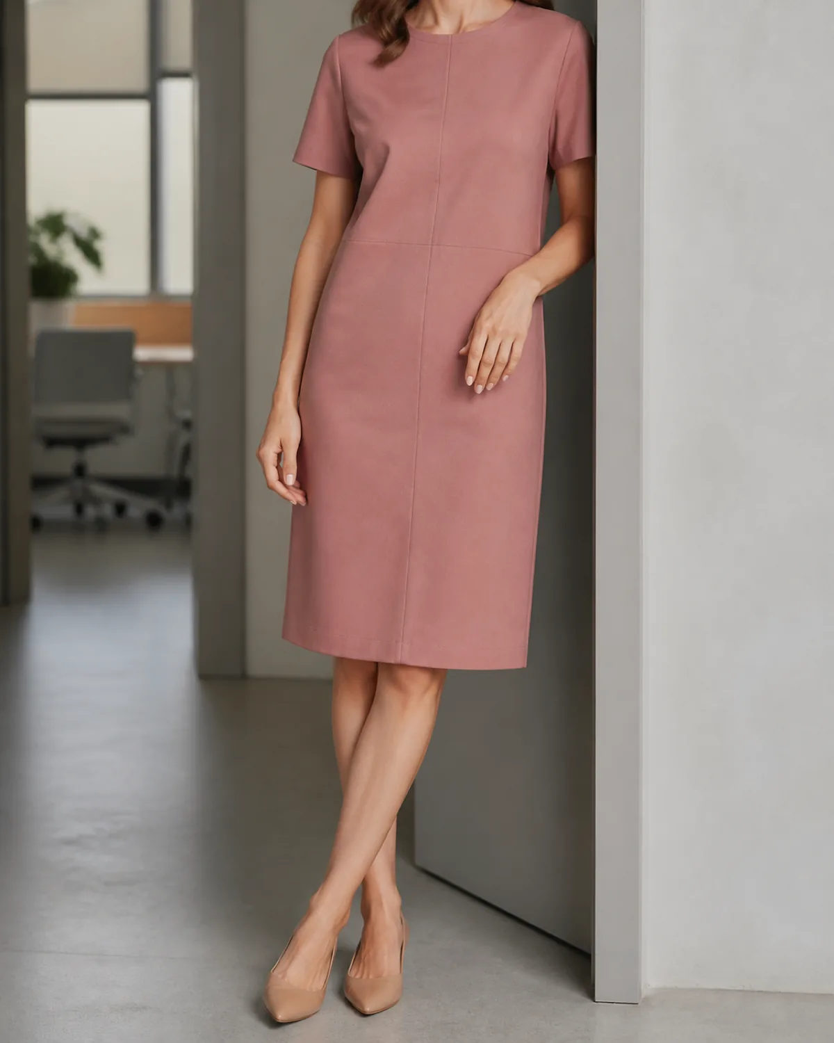 A woman in a dusty-rose crew-neck shift dress standing in a doorway, leaning one shoulder lightly against a light-grey column