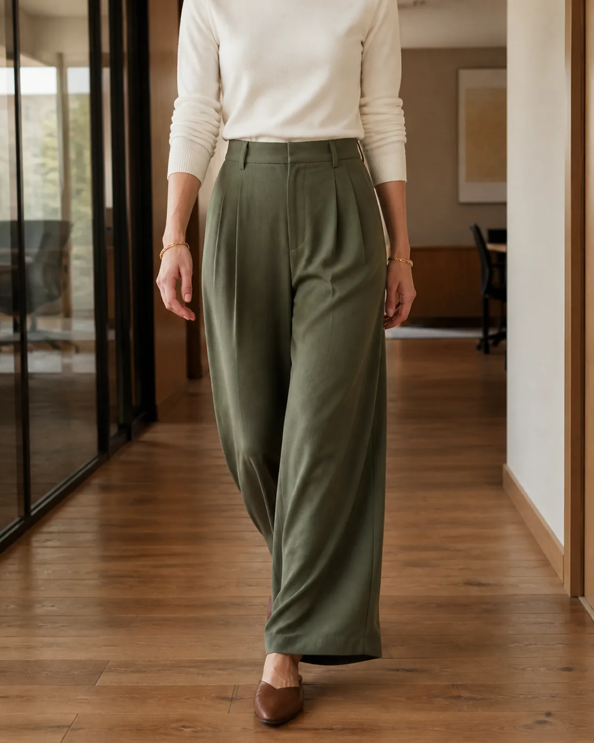 A woman walking mid-stride in olive high-waisted wide-leg trousers with a tucked white crewneck knit top and brown leather mules in an office hallway