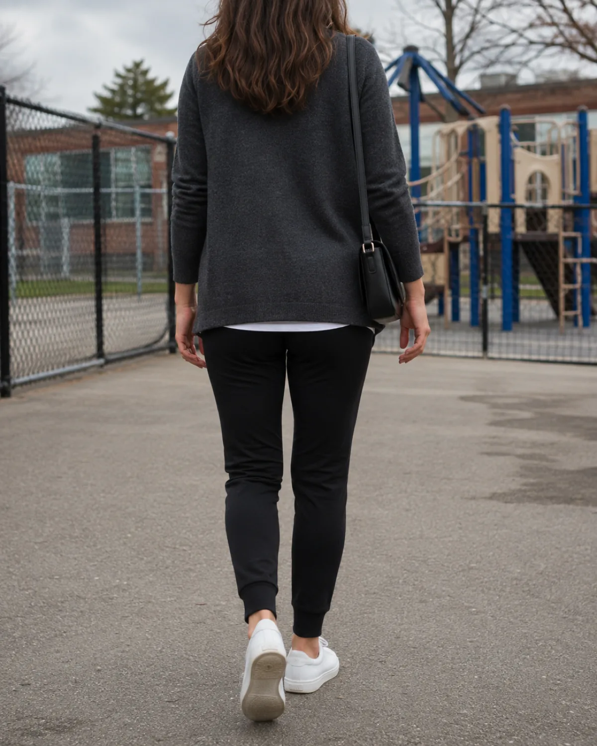 A woman walking away from the camera on a sidewalk near an elementary school playground wearing black ponte joggers, a charcoal structured cardigan, white sneakers, and a tan bag on her shoulder, climbing equipment and a slide visible behind the fence