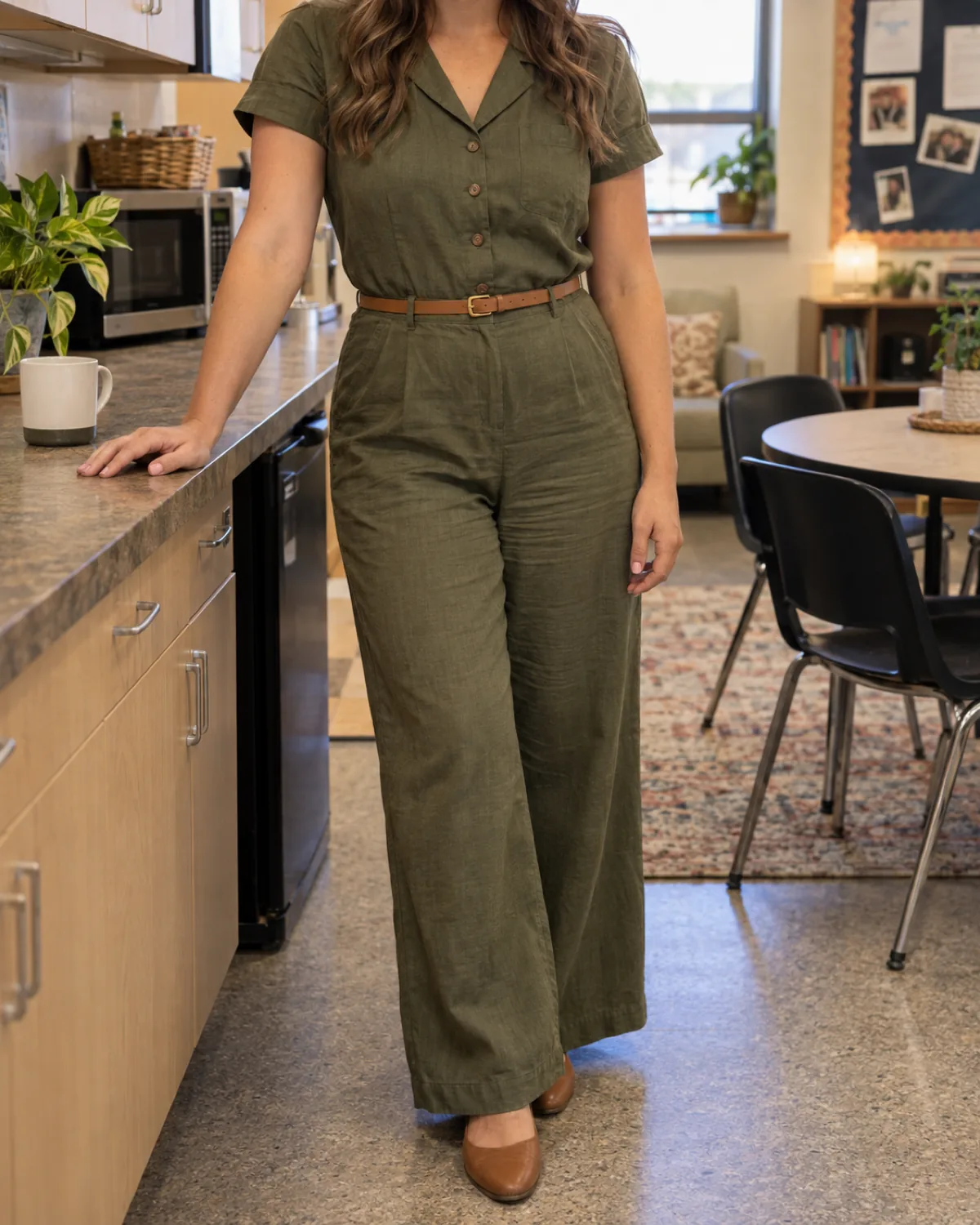 A woman standing in a teacher lounge, one hand on a counter, wearing an olive-green cotton-linen jumpsuit with a notch collar and a thin tan leather belt, a coffee mug and microwave visible behind her