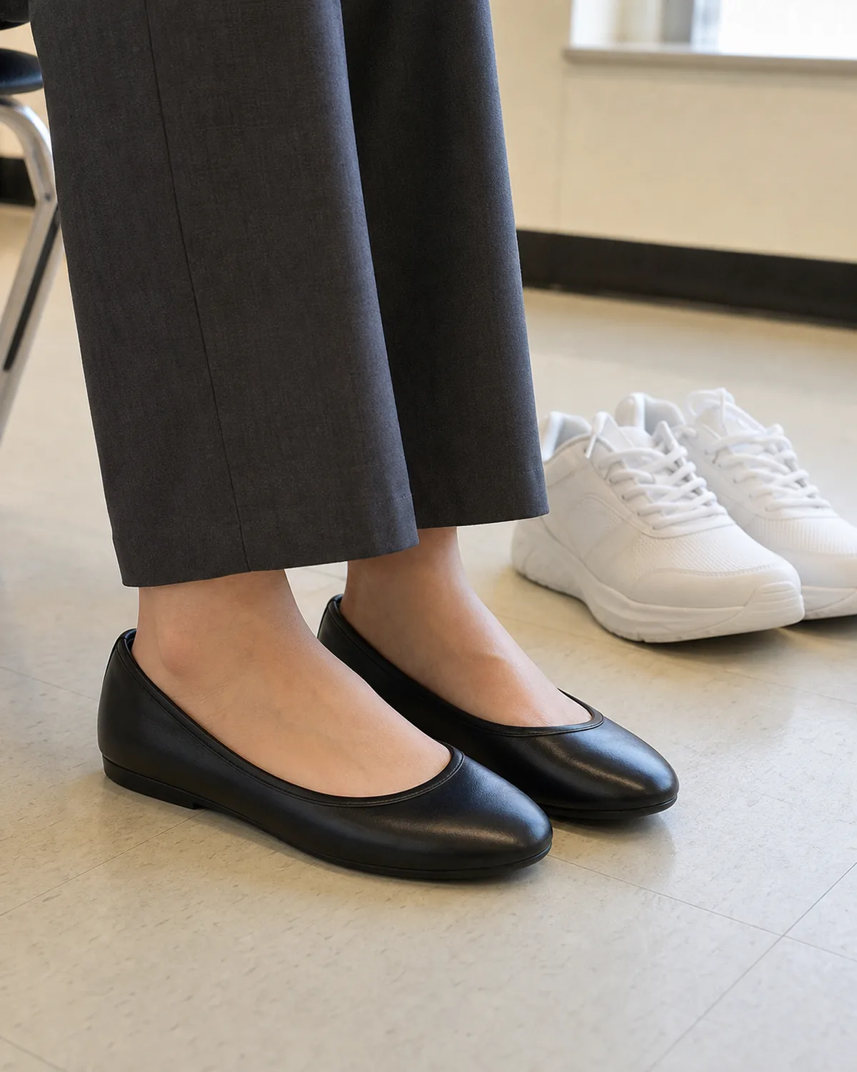 Close crop on a classroom floor showing a woman's feet in black leather closed-toe ballet flats with charcoal trouser hems, and a pair of white athletic sneakers beside them