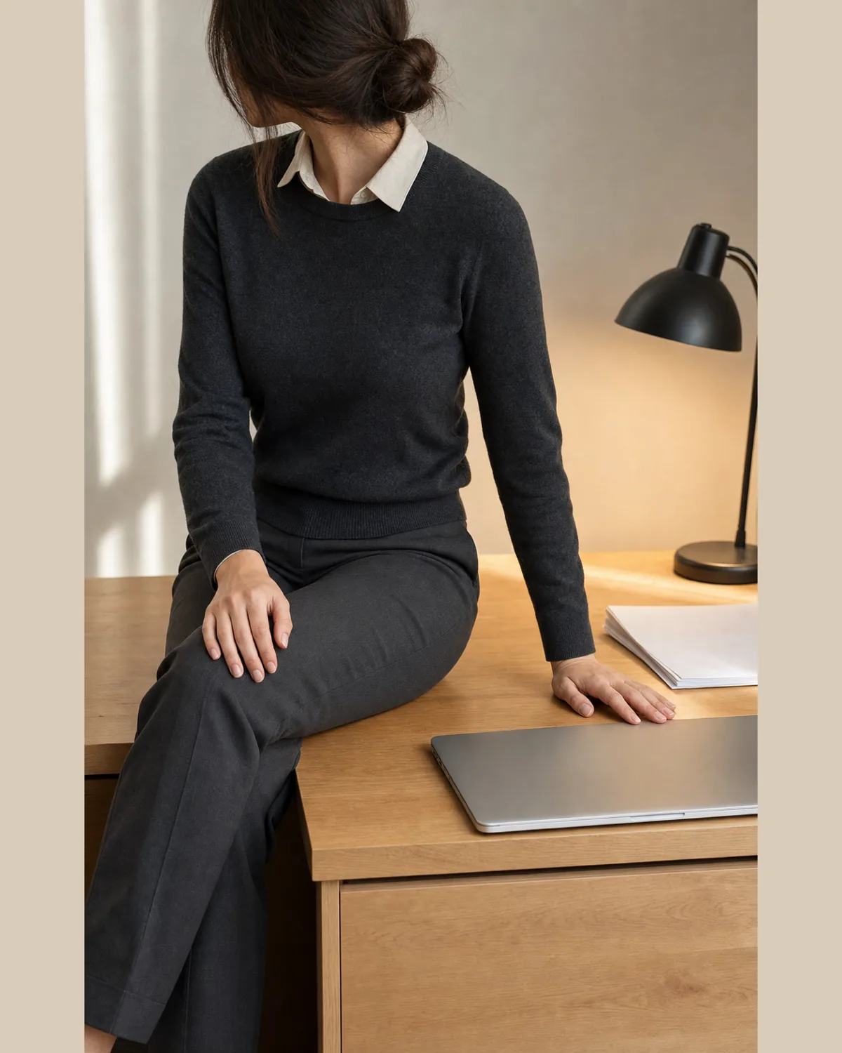 A woman seated at a teacher's desk in a charcoal crewneck knit over an ivory collared shirt and charcoal trousers, with a closed laptop and papers beside her, face turned away