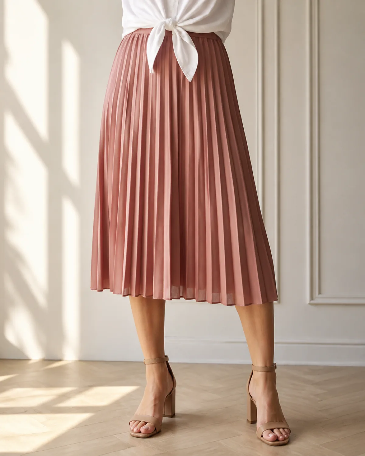 Close crop from the waist down showing a white button-down shirt with tails tied at the front waist over a dusty-rose pleated midi skirt with nude ankle-strap heeled sandals on a pale hardwood floor in warm sidelight