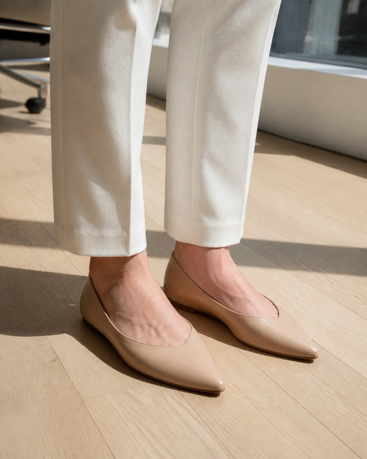 Close crop of nude pointed-toe leather flats on a hardwood office floor with cream trouser hems visible above