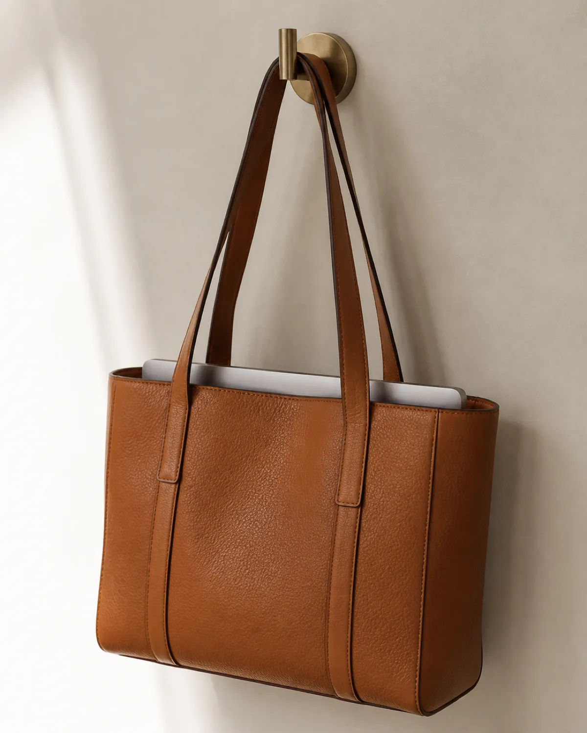 A tan structured leather tote bag hanging from a brass wall hook with a laptop edge visible peeking from the top