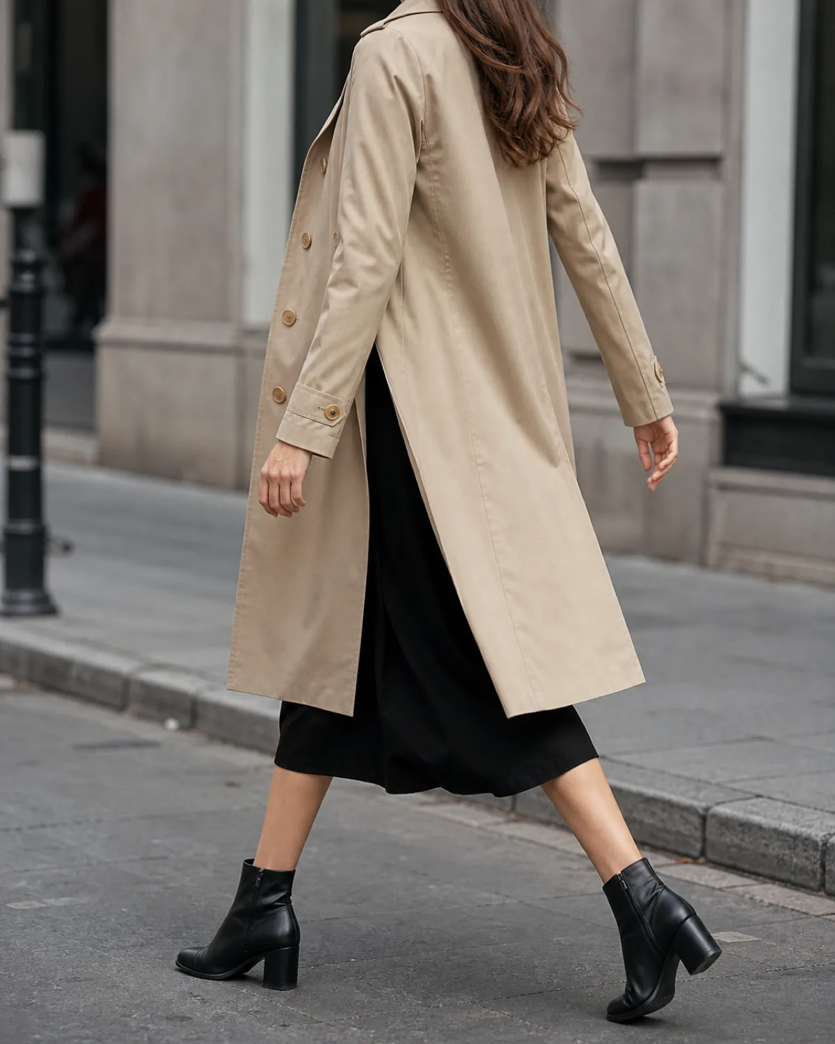 A woman in a black long-sleeve midi dress under an open beige trench coat, walking away from the camera on an urban sidewalk with black leather ankle boots