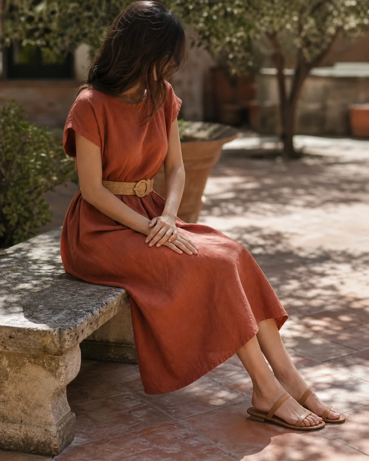 A woman in a terracotta linen-blend midi dress seated on a low stone bench in an outdoor courtyard with a raffia belt and tan leather sandals