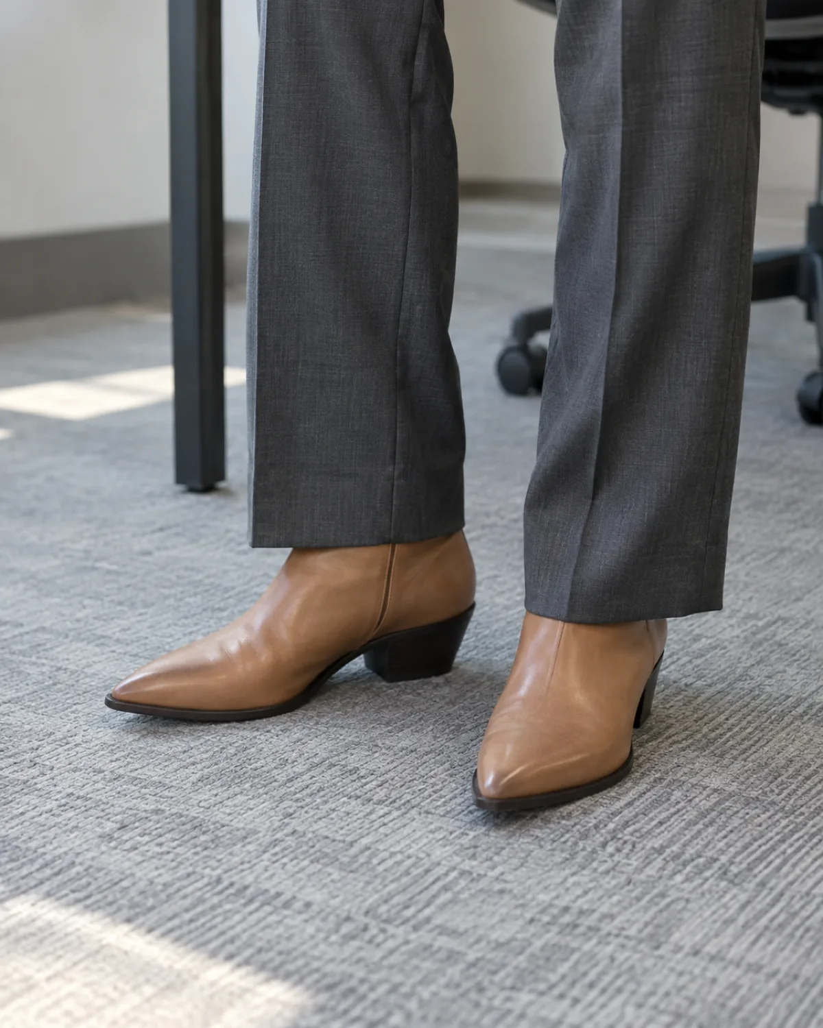 Close crop from mid-shin to floor showing tan leather Western-inflected ankle boots with a pointed toe and low angled heel under charcoal tailored straight-leg trousers on grey office carpet with a desk leg visible in the background