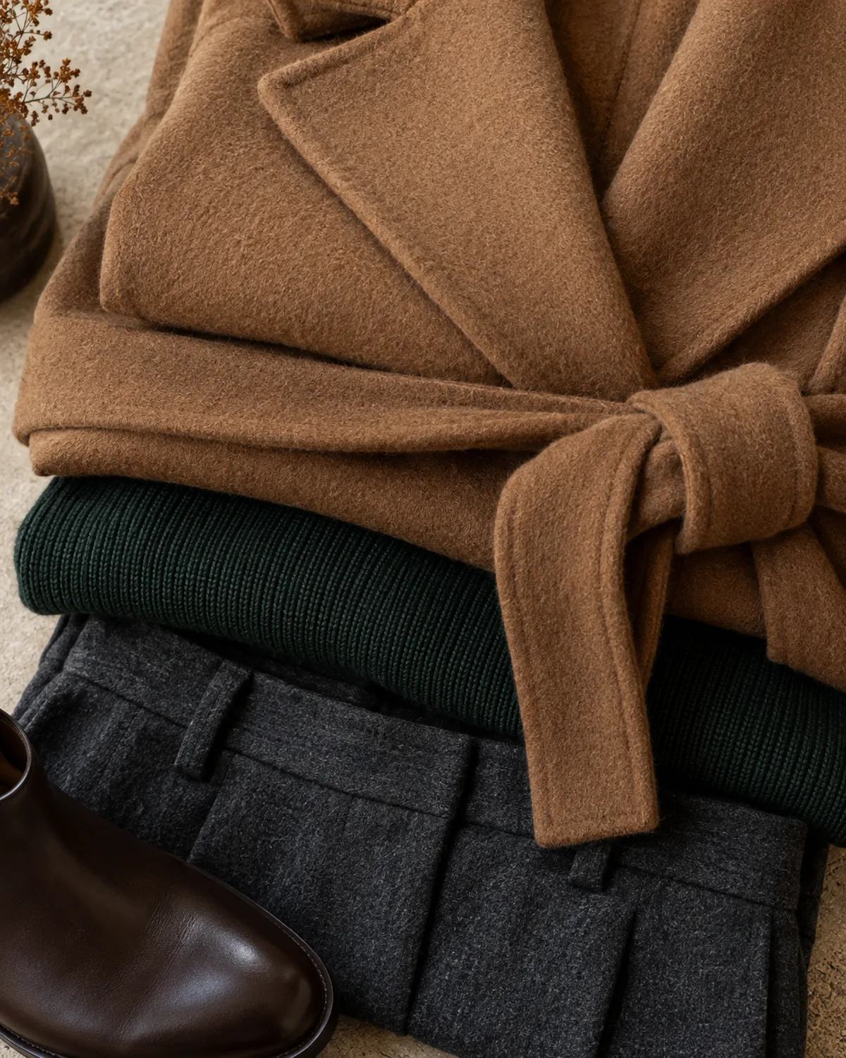 Stacked detail macro showing three fabric layers: a camel wool wrap coat with self-tie belt, a dark green knit, and charcoal high-waisted wool trousers with a brown ankle boot toe visible at the bottom edge