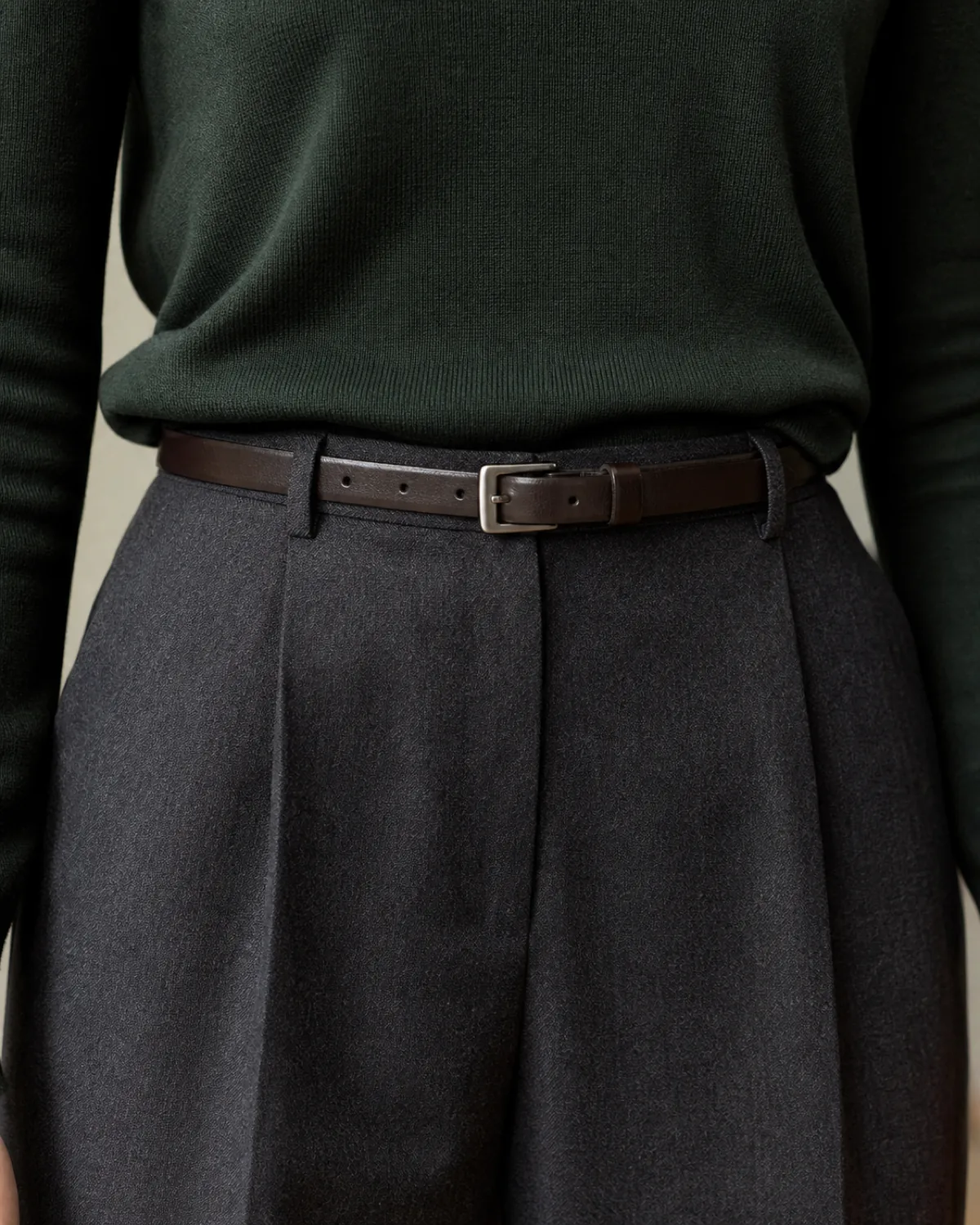 Close crop on the waist showing a forest green fine-knit merino sweater front-tucked into charcoal tapered wool trousers, secured with a thin dark brown leather belt with a small square brushed-metal buckle