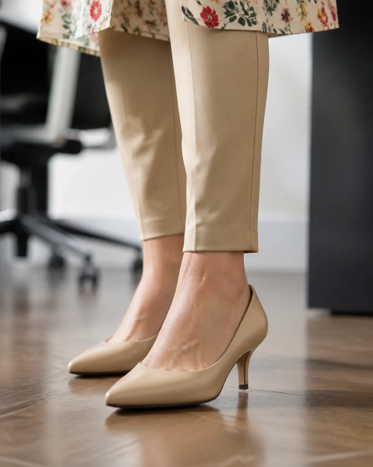 Close-up low-angle shot of slim khaki pants at the ankle with a narrow tapered hem above nude pointed-toe low heels on a hardwood office floor