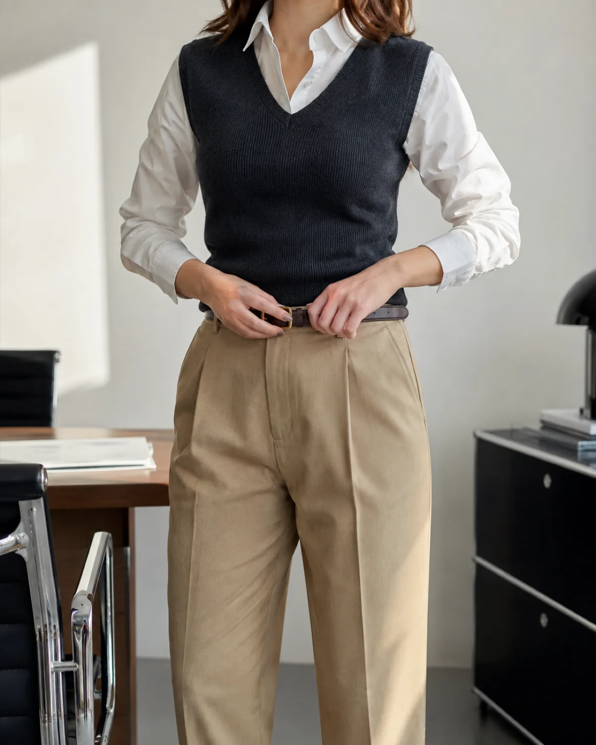 A woman from chin to knee in a modern office wearing high-rise tapered khaki pants, a charcoal knit vest over a white poplin shirt with collar visible, adjusting a dark leather belt with both hands