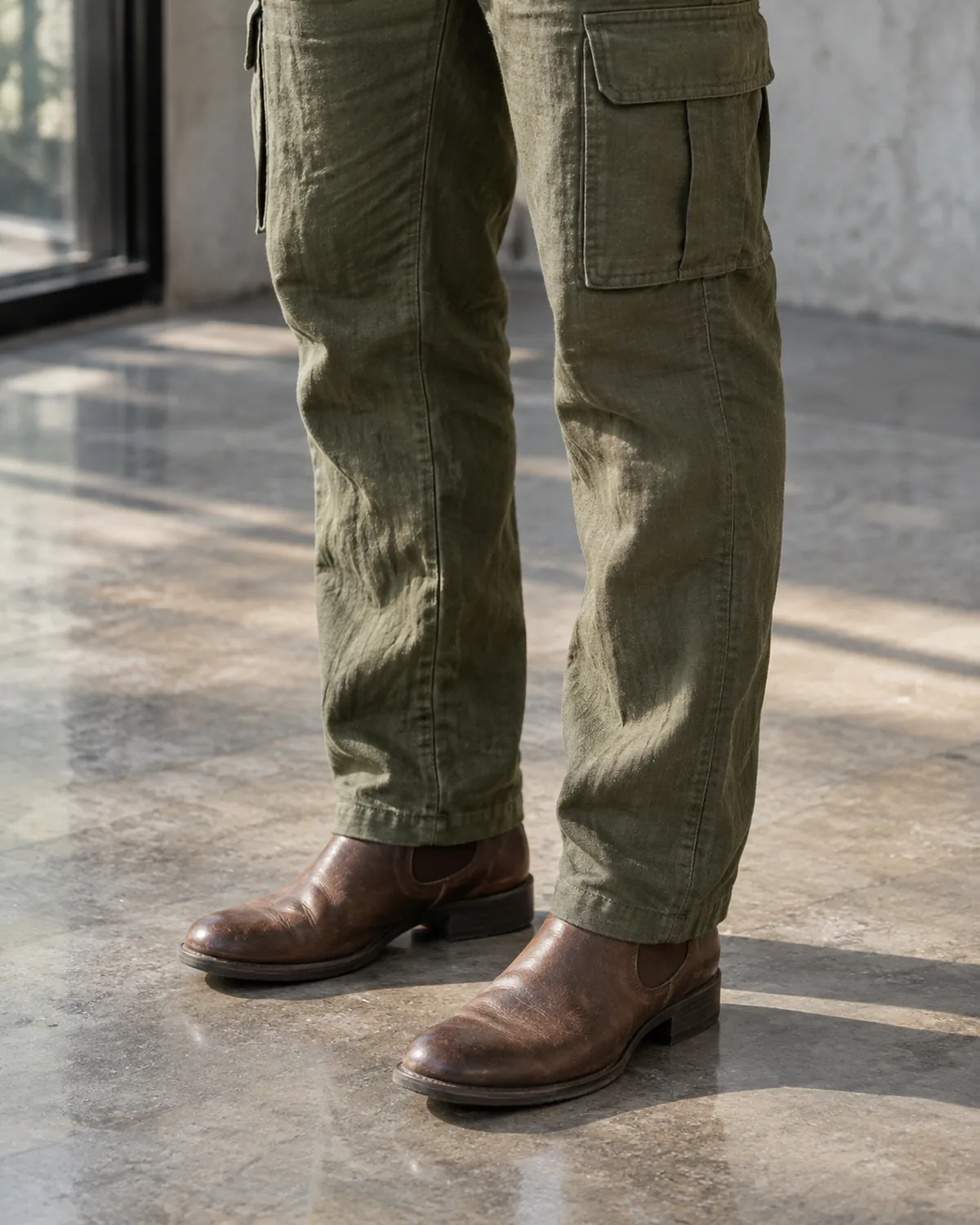 A close crop from the knee to the floor showing olive linen cargo-style trousers with a flat-flap pocket visible on the thigh and brown leather ankle boots on polished concrete