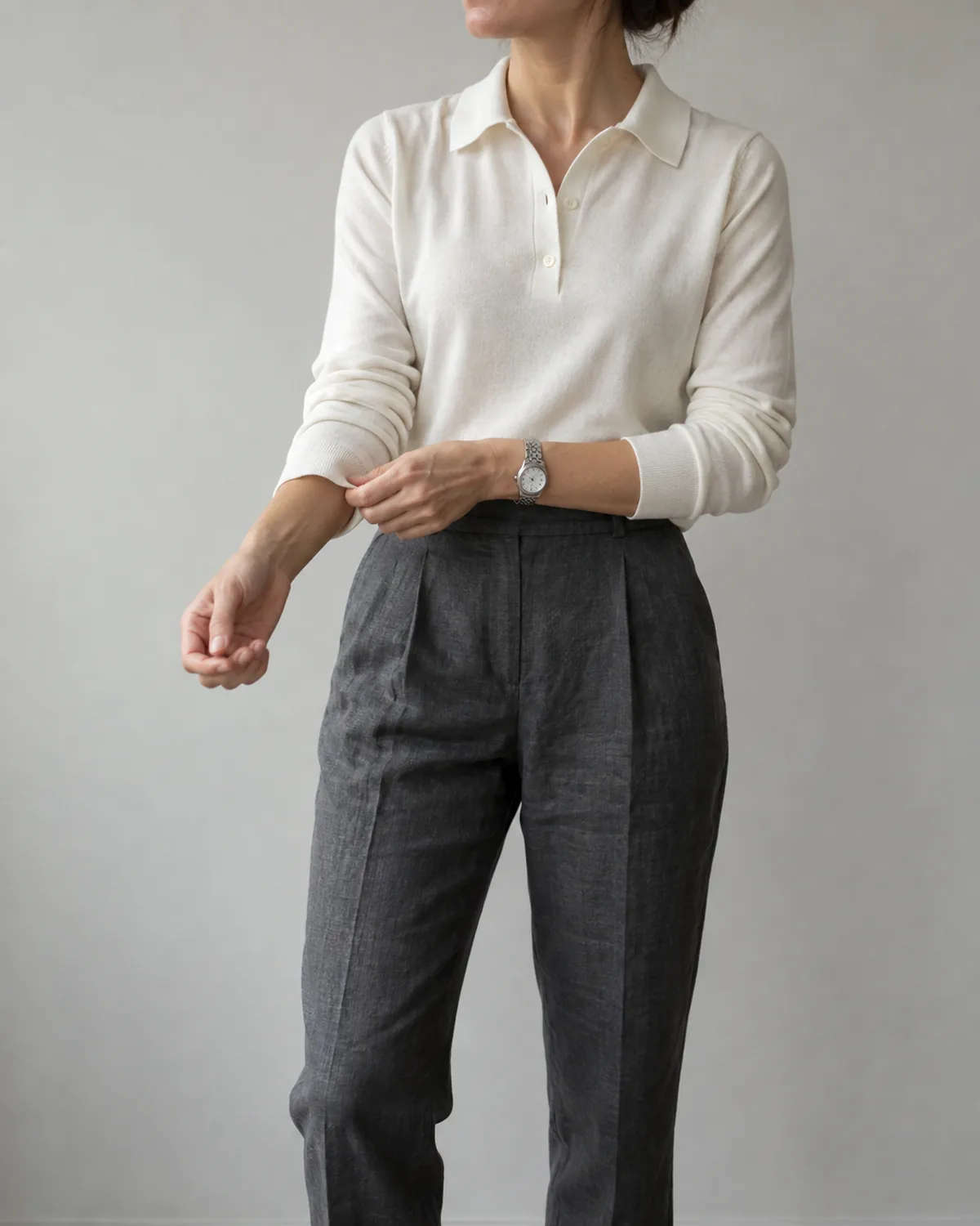 A woman shown from chin to knee adjusting her right shirt cuff, wearing charcoal cropped tapered linen trousers, an ivory silk knit polo with a neat collar, and a silver watch