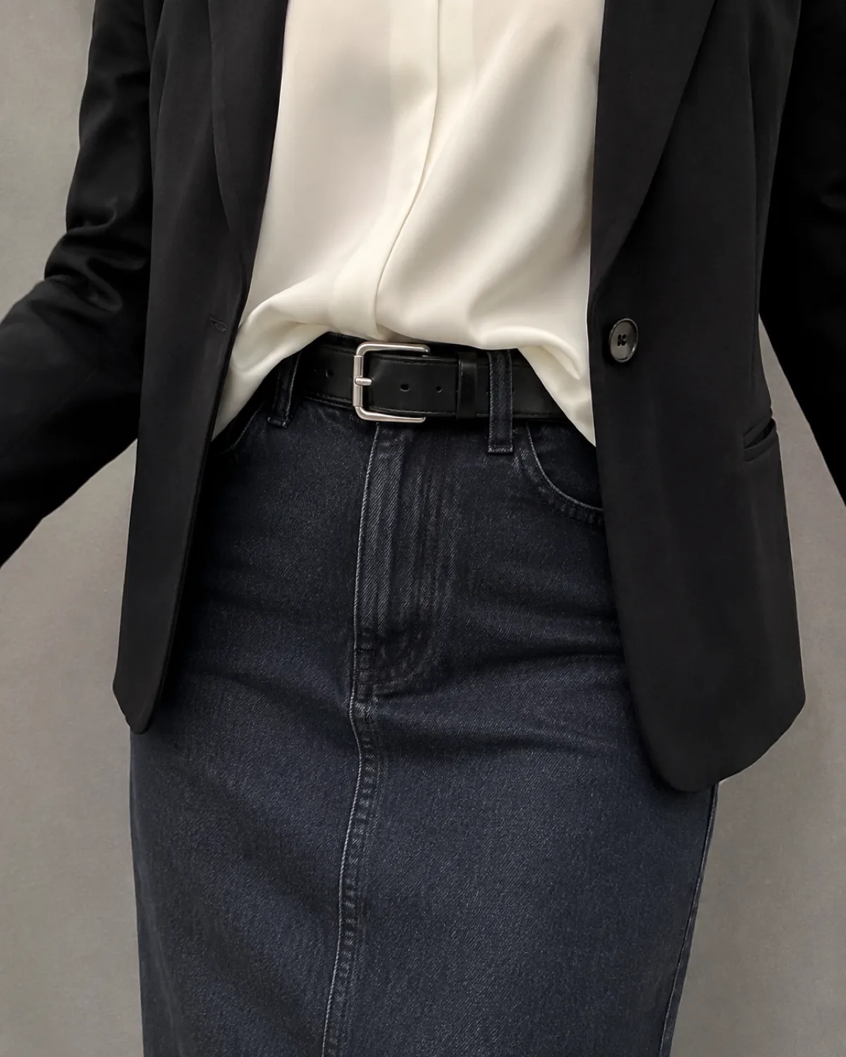 Close crop of a dark-wash denim midi skirt waistband with a black leather belt, white silk blouse French-tucked, and the bottom hem of a black blazer