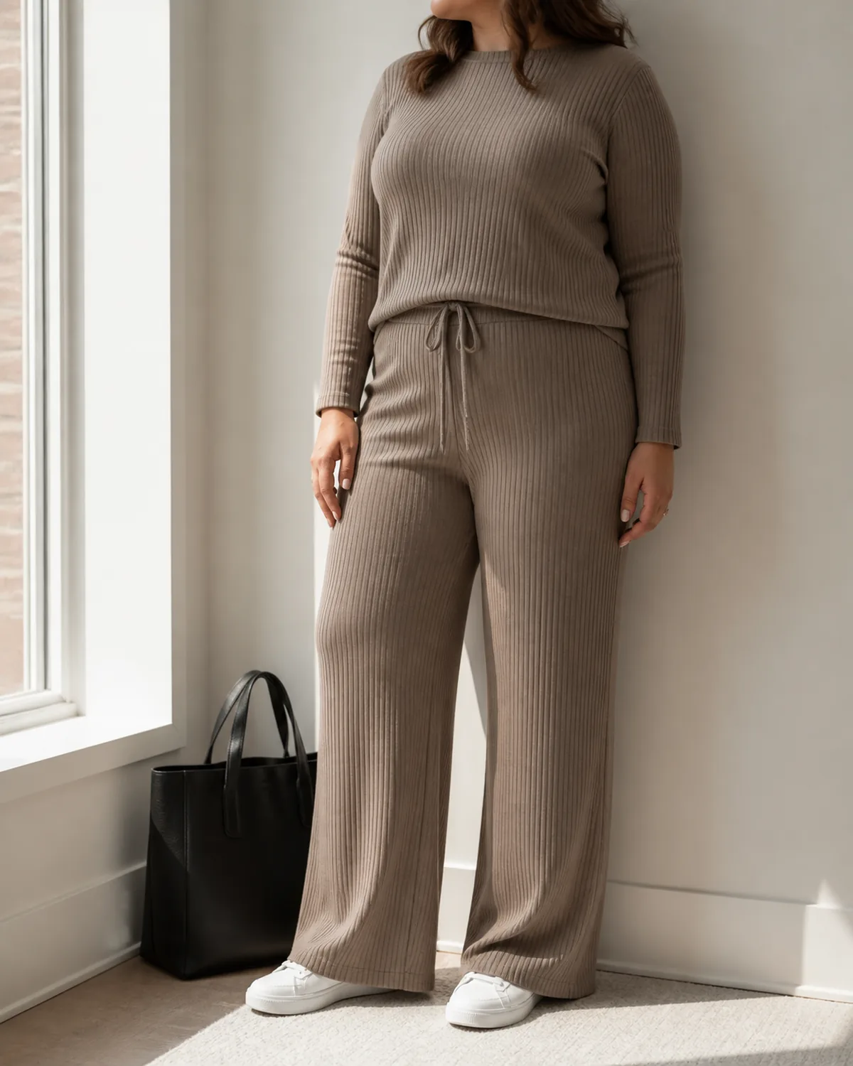 A midsize woman in a taupe ribbed knit co-ord set with wide-leg trousers and a matching crew-neck top, standing near a window with a black leather tote on the floor beside her