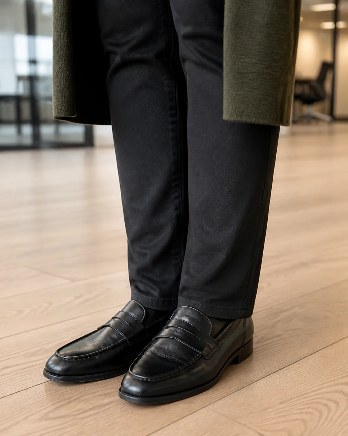 Close crop of slim black ankle pants meeting black leather loafers on a light hardwood office floor with a dark olive long cardigan visible at the edge