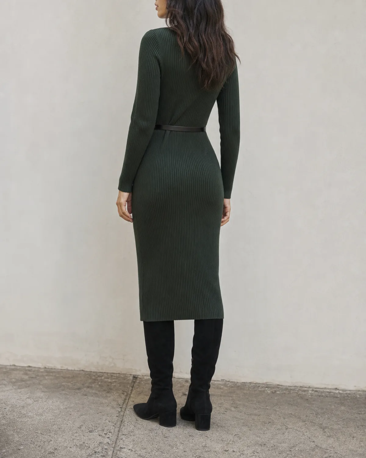 A woman standing at a three-quarter rear angle wearing a dark forest green ribbed knit midi dress with a thin dark belt at the waist and black suede tall boots