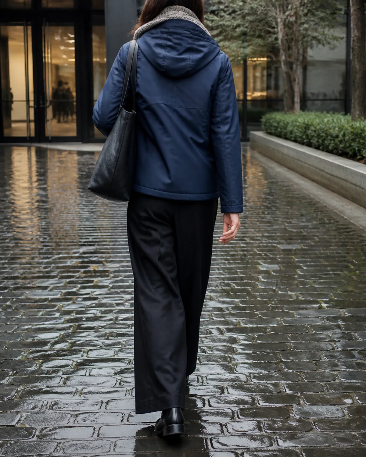 A woman walking away from the camera on wet cobblestones toward an office building entrance, wearing a navy waterproof shell jacket with the hood down, an oatmeal wool scarf at the neck, black wide-leg trousers, and dark leather ankle boots, a black leather tote bag on her shoulder
