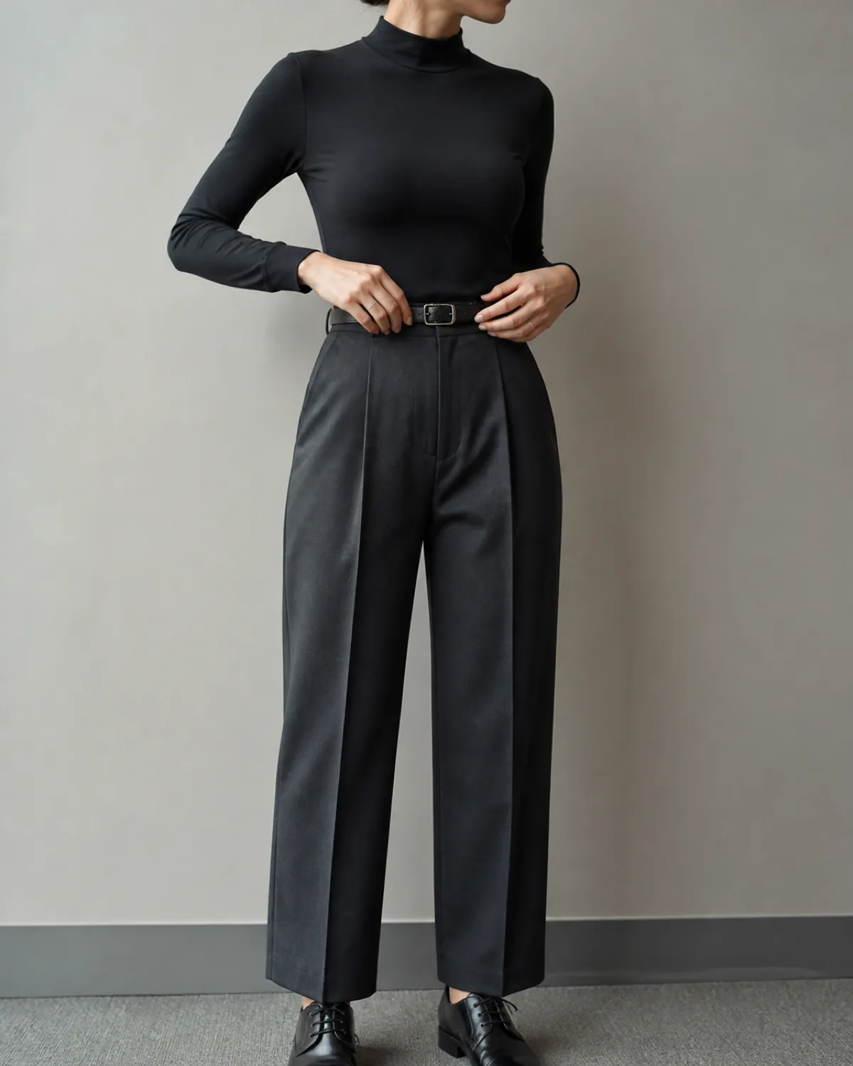 A mid-torso crop of a woman adjusting a thin black leather belt at her waist, wearing charcoal pleated trousers with sharp pressed front pleats and a black mock-neck fitted top with long sleeves, black leather oxford shoes visible at the bottom
