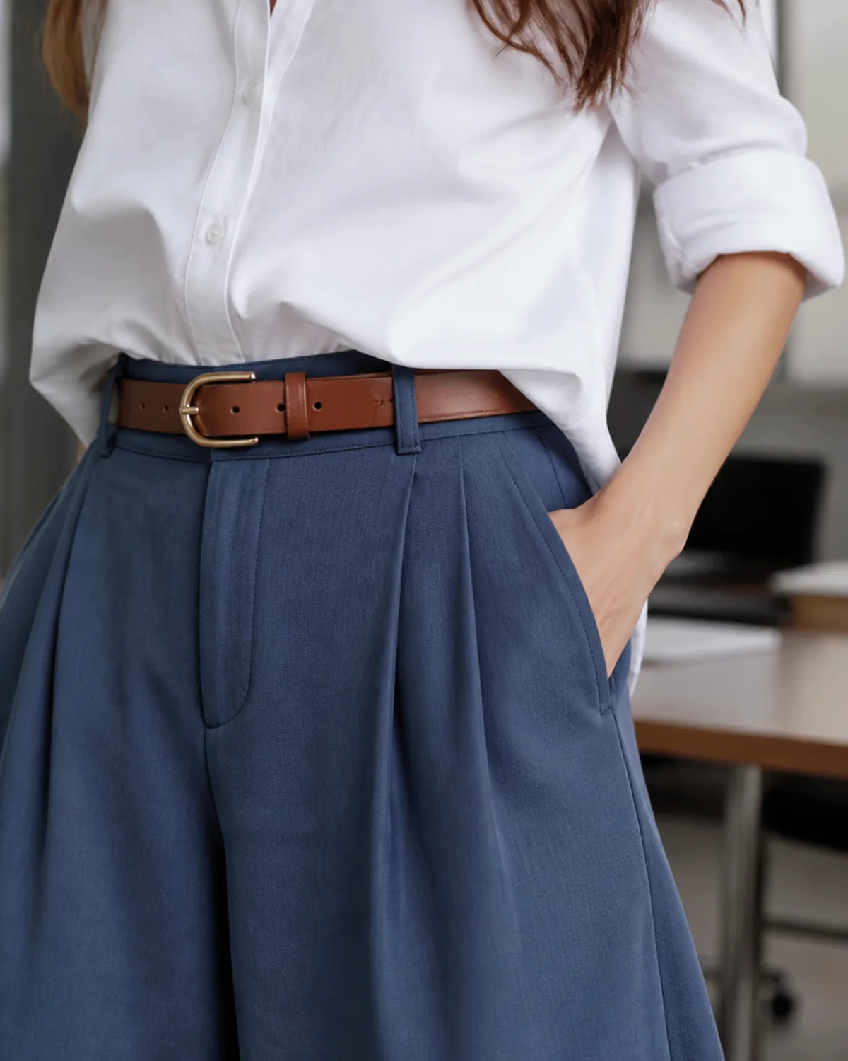 A close-up detail crop centred on the waist area of a woman wearing slate blue wide-leg trousers with a cognac leather belt at the high waist and a white poplin shirt tucked in with a clean French tuck, one hand in the trouser pocket